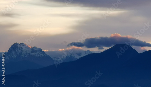 Le cime delle montagne innevate al tramonto nel cielo plumbeo invernale e nuvole bordate di rosso dalla luce del sole