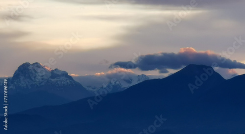 Le cime delle montagne innevate al tramonto nel cielo plumbeo invernale e nuvole bordate di rosso dalla luce del sole © GjGj