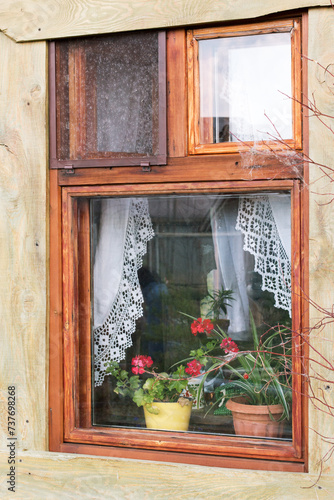 Vintage window in a wooden, rural house. There are flower pots with red flowers and white curtains with lace on the window. Stylization.