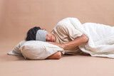 Man in pajamas home, wear sleep mask, lying with pillow and blanket isolated on beige background