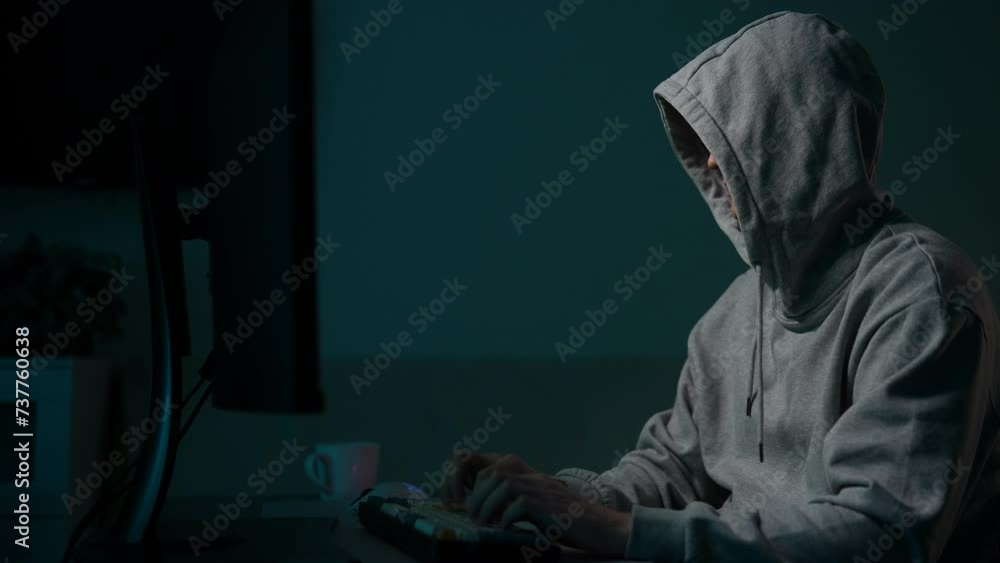 Focused unrecognizable man hacker wearing sweatshirt with hood typing on desktop computer keyboard breaking password. Dangerous hooded computer hacker infecting system with cyber virus in dark room. Focused unrecognizable man hacker wearing sweatshirt with hood typing on desktop computer keyboard breaking password. Dangerous hooded computer hacker infecting system with cyber virus in dark room.
