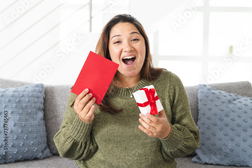 Excited young biracial woman at home, holding a gift and a card on a video call