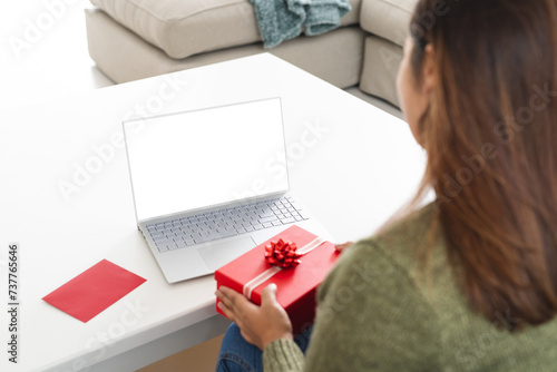 Young biracial woman at home, holding a gift with a laptop on the table, on a video call