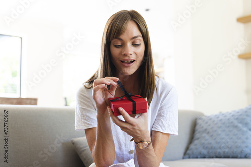 A young Caucasian woman appears pleasantly surprised as she opens a small red gift box