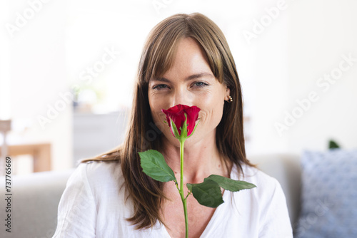 A middle-aged Caucasian woman smiles while holding a red rose close to her face