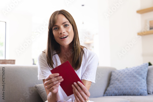 A young Caucasian woman smiles, holding a red book in a bright room, on a video call
