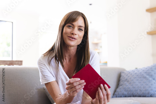 A young Caucasian woman smiles at the camera, holding a red book on a video call