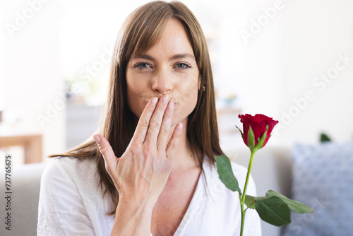 A young Caucasian woman blows a kiss towards the camera, holding a red rose in her hand