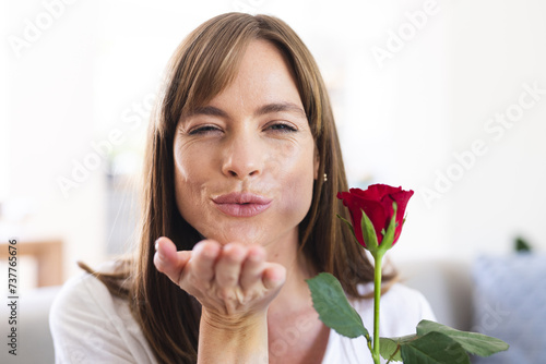A middle-aged Caucasian woman is playfully blowing a kiss while holding a red rose