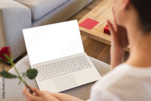 A young woman is seen from behind holding a rose while looking at a laptop screen, with copy space