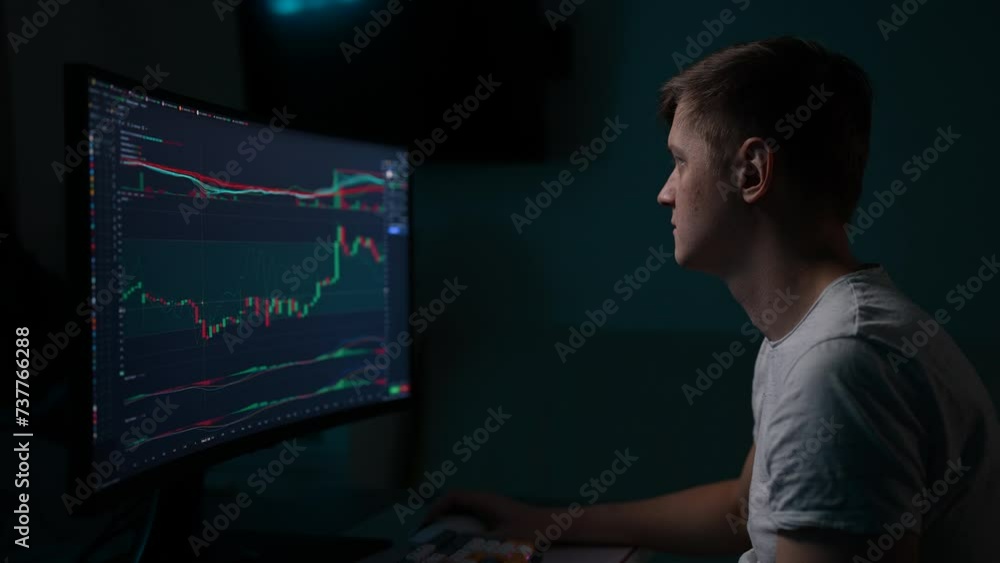 Pensive male stock trader watching real-time stocks, exchange market charts on desktop monitor sitting at desk in dark room. Man works remotely in investment at home office. Shooting in slow motion. Pensive male stock trader watching real-time stocks, exchange market charts on desktop monitor sitting at desk in dark room. Man works remotely in investment at home office. Shooting in slow motion.