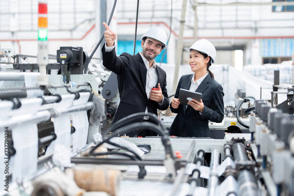 Asian female engineer and European male operator of an industrial plant discuss work and point fingers at steel and plastic production machinery, have laptops and walkie-talkies Wear a suit and helmet
