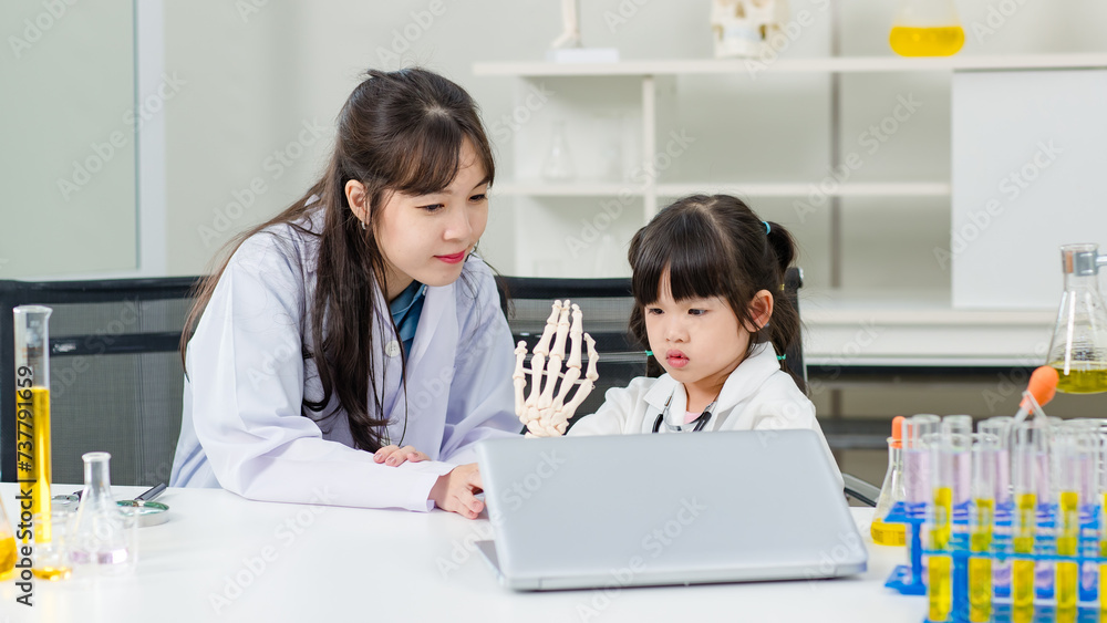 Fototapeta premium Asian woman doctor physiotherapist explains human fake body skeleton model on a table to little children girl at laboratory study room. Education anatomical human concept learning for kids.