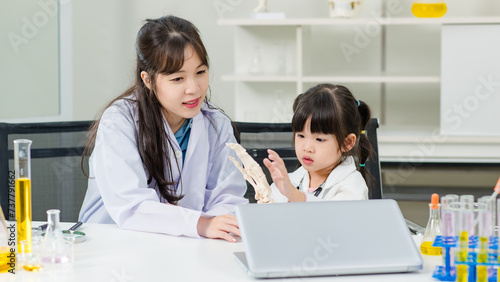 Asian woman doctor physiotherapist explains human fake body skeleton model on a table to little children girl at laboratory study room. Education anatomical human concept learning for kids.