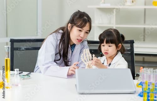 Asian woman doctor physiotherapist explains human fake body skeleton model on a table to little children girl at laboratory study room. Education anatomical human concept learning for kids.