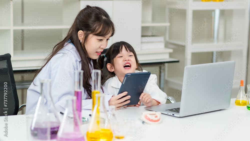 Obraz premium Asian woman scientist and little children girl reading laptop and tablet computer for data learning at chemical laboratory study room. Education research and development concept learning for kids.