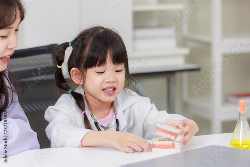 Asian woman doctor physiotherapist explains human fake skeleton teeth model on a table to little cute children girl at laboratory study room. Education anatomical human concept learning for kids.