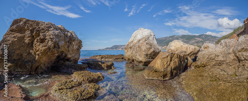 Panoramic view of the rocky coast of the Mediterranean Sea with crystal clear water and between the stones you can see the silhouette in the distance of a sailing ship with lowered sails in Alanya.