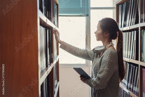 Young asian female college student taking book from shelf in library.
