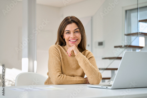 Happy smiling older mature middle aged 45 years old woman using laptop computer sitting at table, hybrid working, elearning or relaxing with device at home, looking at camera, portrait. photo