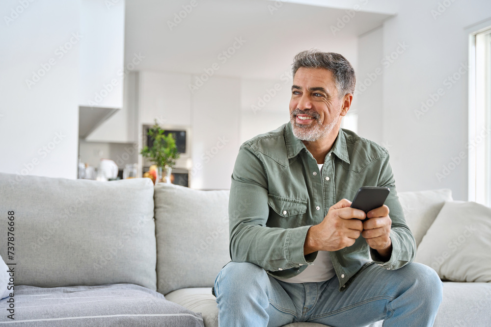 Happy older mature middle aged man holding cell phone using smartphone sitting at home on sofa, relaxing on couch in living room, looking away at copy space advertising mobile offers and apps.