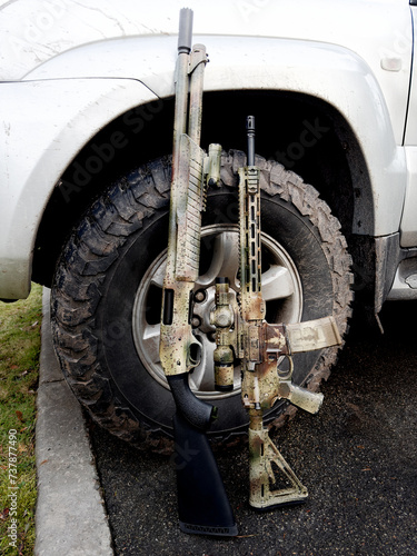 A shotgun and a rifle gun standing against the wheel of a silver SUV photo