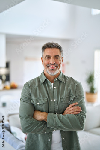 Happy confident middle aged senior man standing with arms crossed at home. Smiling older mature 50 years old handsome man looking at camera posing in modern house living room. Vertical portrait.