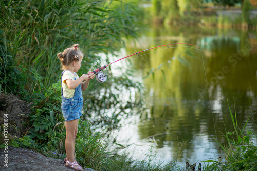 Three-year-old blond girl in denim shorts with straps catches fish on the river with a black and white cat