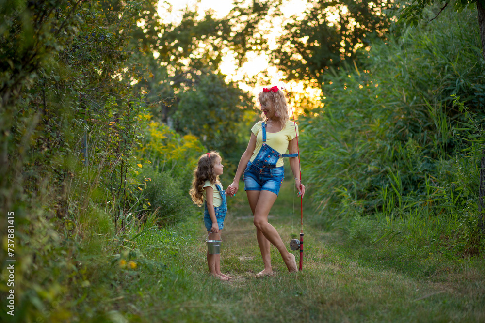 Two blond barefoot fisherwomen in denim shorts with straps with a fishing rod and bucket on a road surrounded by greenery against the backdrop of the setting sun