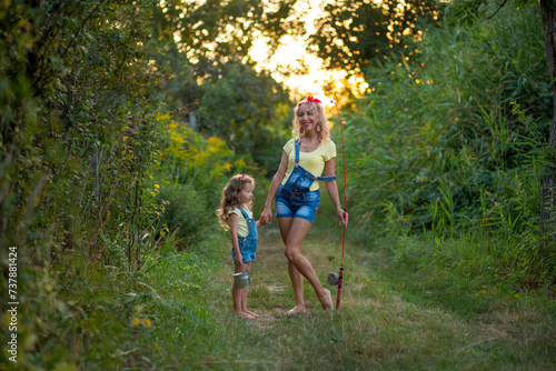 Two blond barefoot fisherwomen in denim shorts with straps with a fishing rod and bucket on a road surrounded by greenery against the backdrop of the setting sun