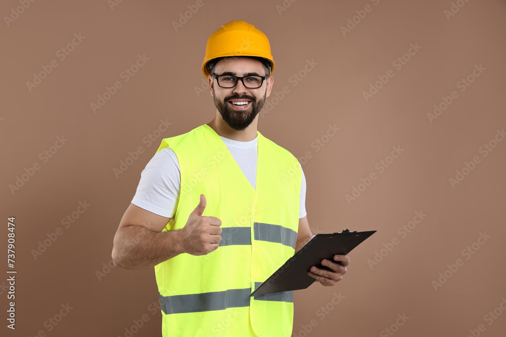 Engineer in hard hat holding clipboard and showing thumb up on brown background Engineer in hard hat holding clipboard and showing thumb up on brown background