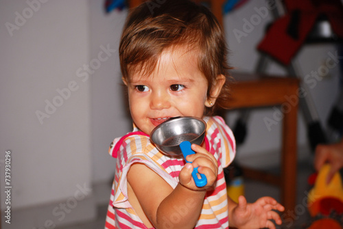 A charming half-length portrait capturing a smiling little girl dressed in a cheerful striped outfit. The little girl gazes forward, playfully chomping once again on a metal colander. This playful sce photo