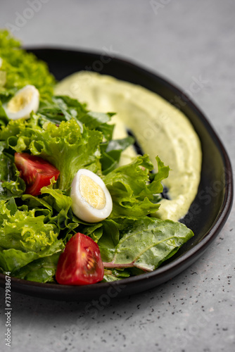 Fresh green salad with quail eggs, cherry tomatoes and guacamole in black plate on grey background