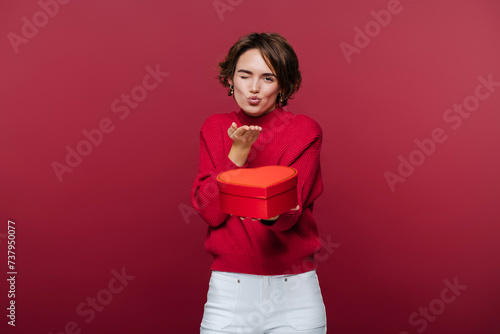 Cute french beautiful young woman smiles at camera, sending air kiss, while holding red gift box