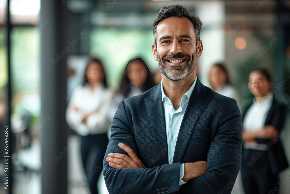 Confident businessman leader with crossed arms and blurred collaborative team on modern office background Confident businessman leader with crossed arms and blurred collaborative team on modern office background
