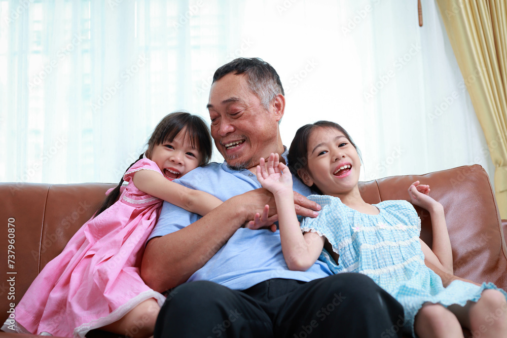 Grandfather and two cute little granddaughter's sitting on the sofa in the house, smiling, laughing, talking and having fun. happy time Love and warmth. Asian family concept Grandfather and two cute little granddaughter's sitting on the sofa in the house, smiling, laughing, talking and having fun. happy time Love and warmth. Asian family concept