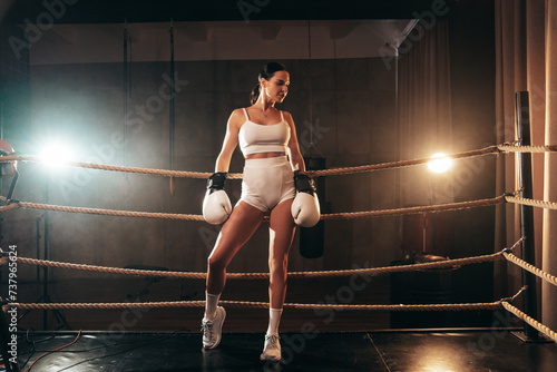 Cinematic portrait of sexy fighter woman in a boxing ring. Model wearing white gloves and braided hair. Hot confident female relax after training for fight match. Stands near the rope. In a smoke