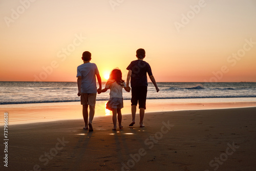 Three kids silhouettes running and jumping on beach at sunset. happy family, two school boys and one little preschool girl. Siblings having fun together. Bonding and family vacation. photo