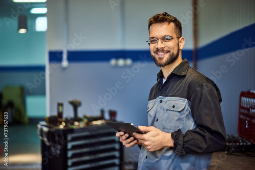 Portrait of a smiling mechanic, posing for the camera with a tablet in his hands, at his workplace, auto service.