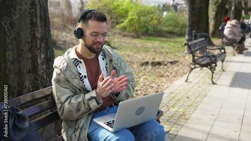 Young businessman working online having video call on laptop sitting on bench outside of office building. Independent expat freelance hipster computer programmer expert programming while traveling