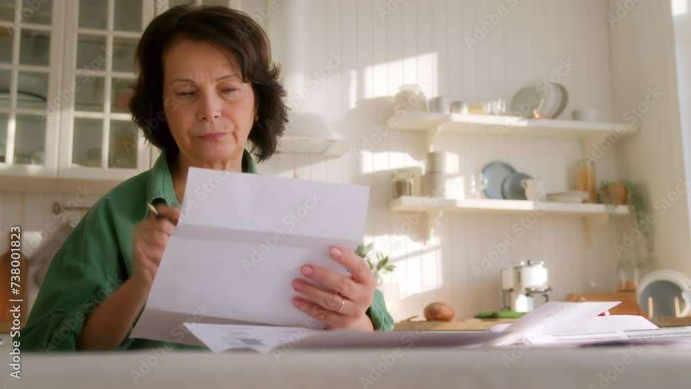 elderly dark haired woman sits at table with papers alone in kitchen in morning. grandma researchers carefully, reads and takes notes in bills, analyzing and planning budget and finance elderly dark haired woman sits at table with papers alone in kitchen in morning. grandma researchers carefully, reads and takes notes in bills, analyzing and planning budget and finance