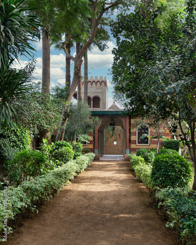 A pathway winds through lush greenery, leading to a Mamluk style building nestled among trees and bushes, creating a serene landscape with a mix of plants and fixtures A pathway winds through lush greenery, leading to a Mamluk style building nestled among trees and bushes, creating a serene landscape with a mix of plants and fixtures