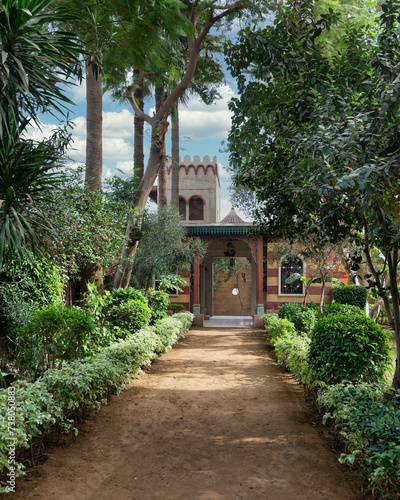 A pathway winds through lush greenery  leading to a Mamluk style building nestled among trees and bushes  creating a serene landscape with a mix of plants and fixtures