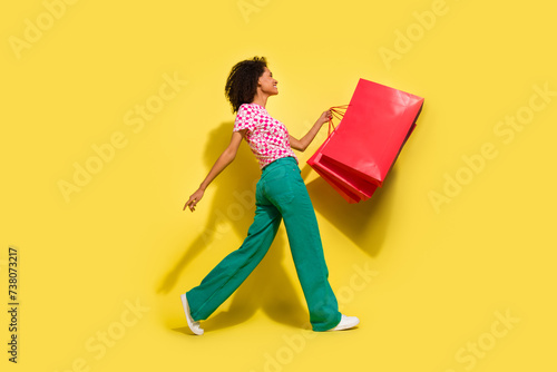Full size profile photo of lovely positive girl hold store bags walking isolated on yellow color background