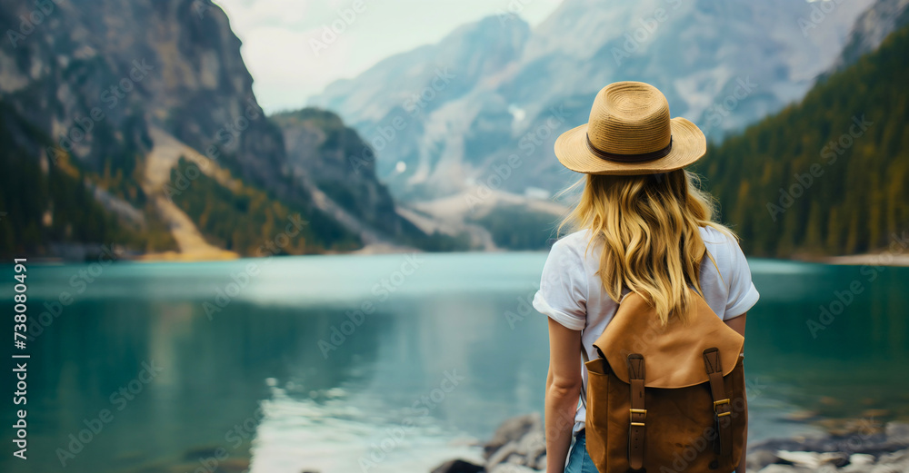 Rearview photography of a young woman with blonde hair wearing a white t shirt, straw hat and a backpack. Standing on a rocky ground in nature and looking at the lake and mountain landscape outdoors
