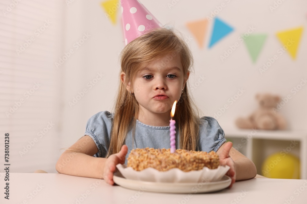 Cute girl in party hat with birthday cake at table indoors Cute girl in party hat with birthday cake at table indoors