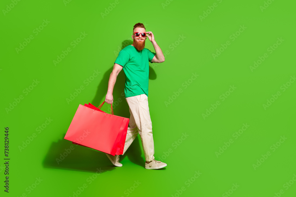 Full body profile portrait of cheerful nice man hold mall bags walk empty space isolated on green color background Full body profile portrait of cheerful nice man hold mall bags walk empty space isolated on green color background