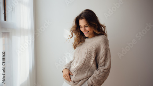A smiling pregnant woman wearing a cozy knit sweater, cradling her belly with love and anticipation, against a minimalist white wall. photo