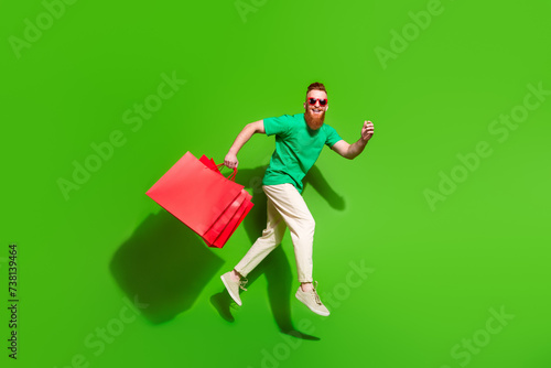 Full body profile photo of crazy cheerful man jump run hold mall bags isolated on green color background