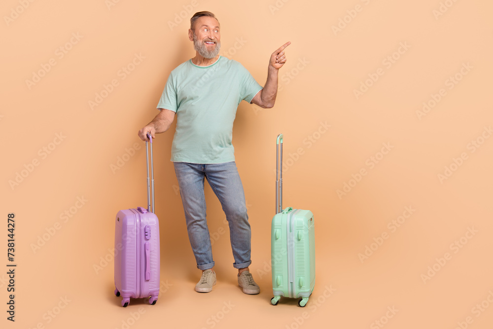 Full length photo of positive aged man hold suitcase look direct finger empty space offer isolated on beige color background Full length photo of positive aged man hold suitcase look direct finger empty space offer isolated on beige color background